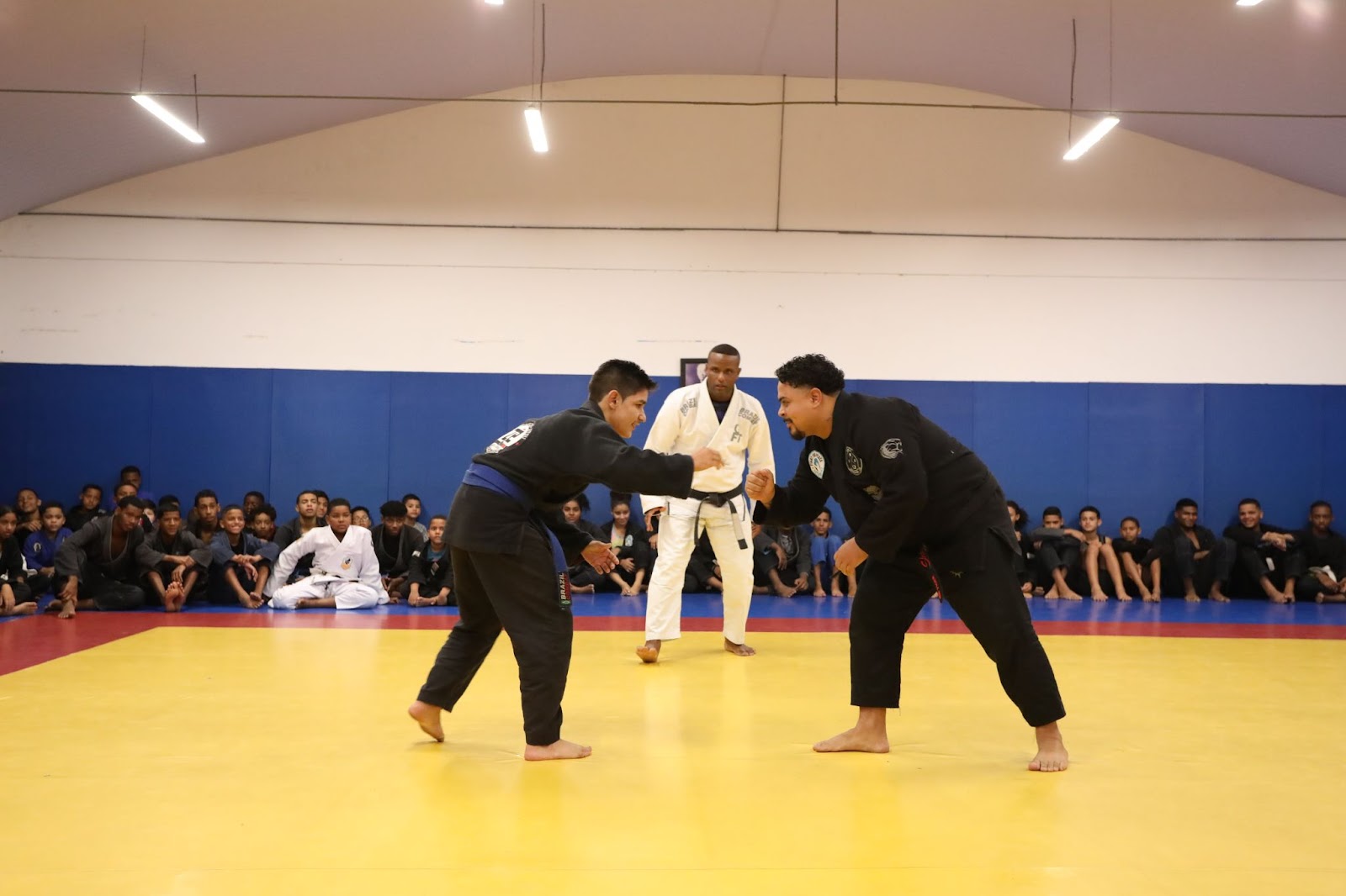 A imagem retrata uma aula de Capoeira, com um grupo de crianças e jovens praticando. Dois participantes estão no centro da ação, realizando movimentos característicos da luta, com expressões de concentração. Todos estão vestidos com uniformes brancos, que contrastam com o piso cinza do local. Ao fundo, há instrumentos musicais típicos da Capoeira, como um berimbau e um pandeiro. O espaço é bem iluminado, com espelhos refletindo os movimentos dos praticantes. A atmosfera é vibrante.  