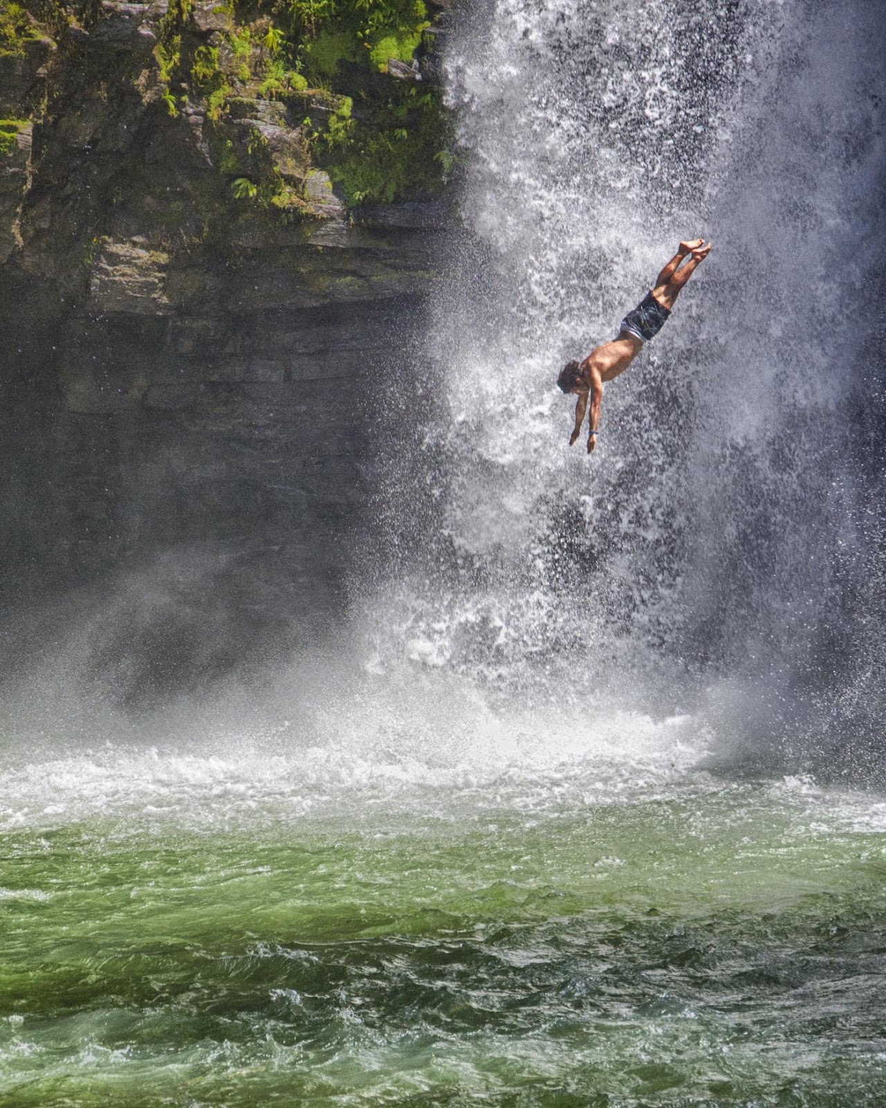 A man is jumping in the water from waterfall and in surrounding there is a large rock