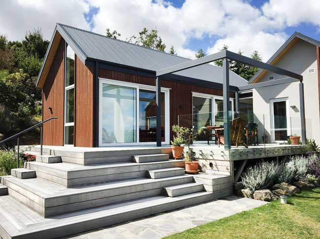 A modern house with wooden siding features large uPVC windows and a glass-railed deck. Surrounded by greenery, it boasts a charming set of steps leading to the entrance.