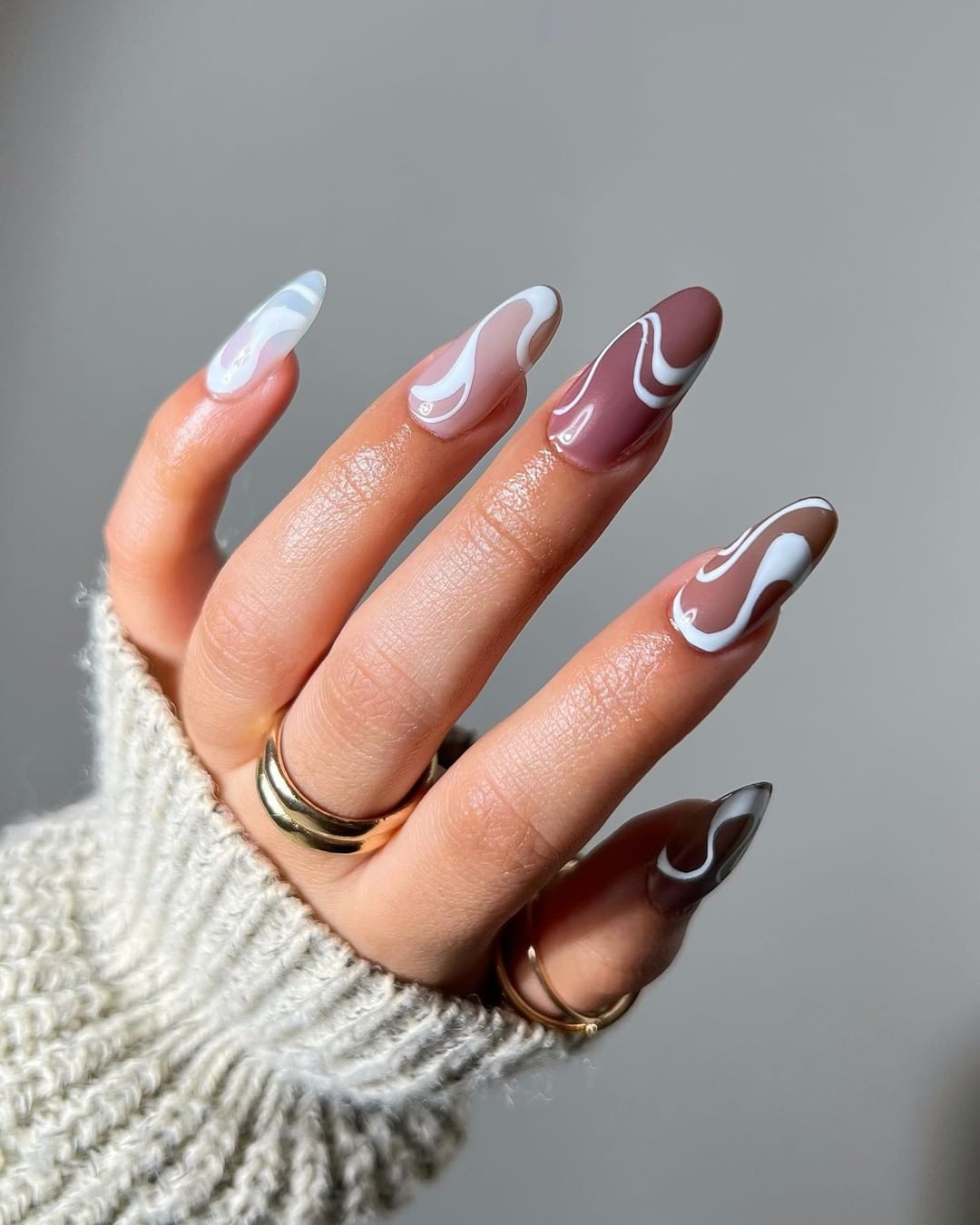 Close-up of a hand with long, almond-shaped nails featuring a glossy brown and white swirl design.

