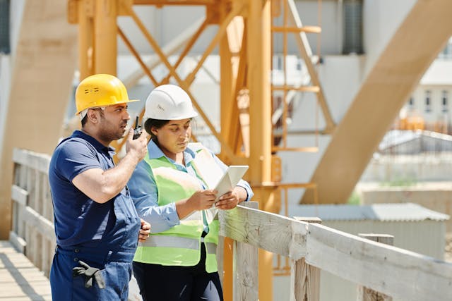 Two construction workers, one in a yellow hard hat and the other in a white hard hat and safety vest, stand on a bridge consulting a tablet as they work with an architectural firm.