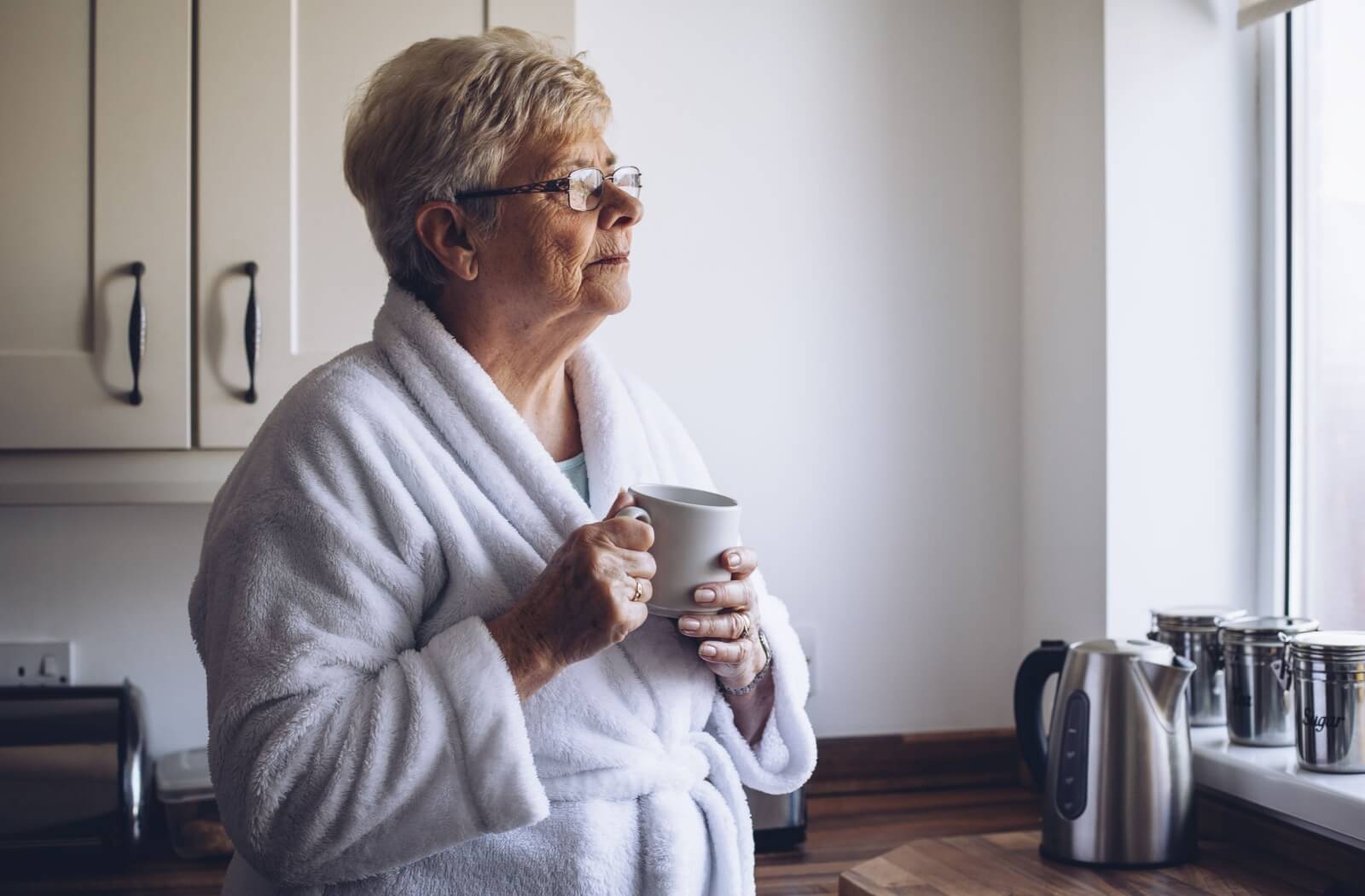 A lone older adult woman in her robes looking out the window while holding a cup.