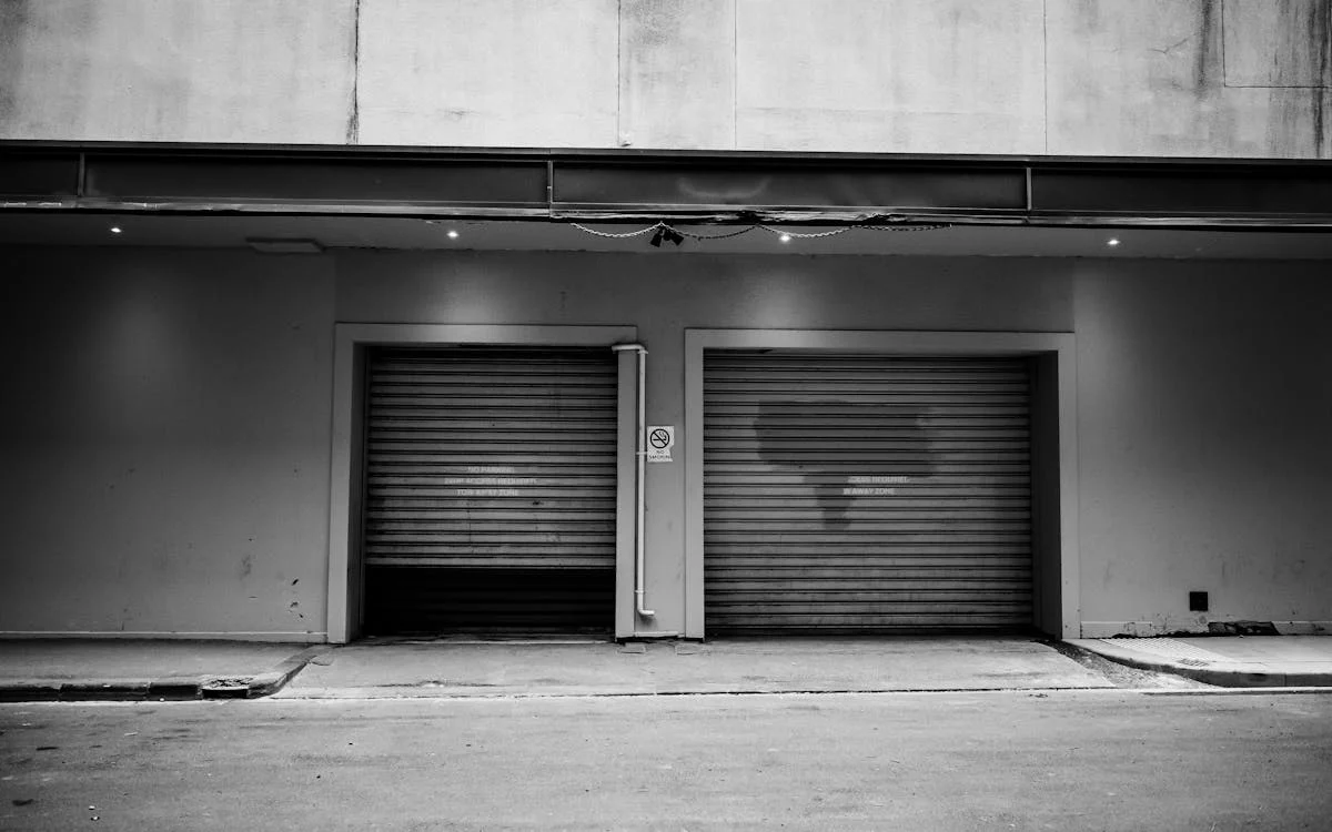 A black-and-white two closed industrial roller doors on a plain building, with a no-smoking sign and dim overhead lighting, creating an urban, minimalist atmosphere