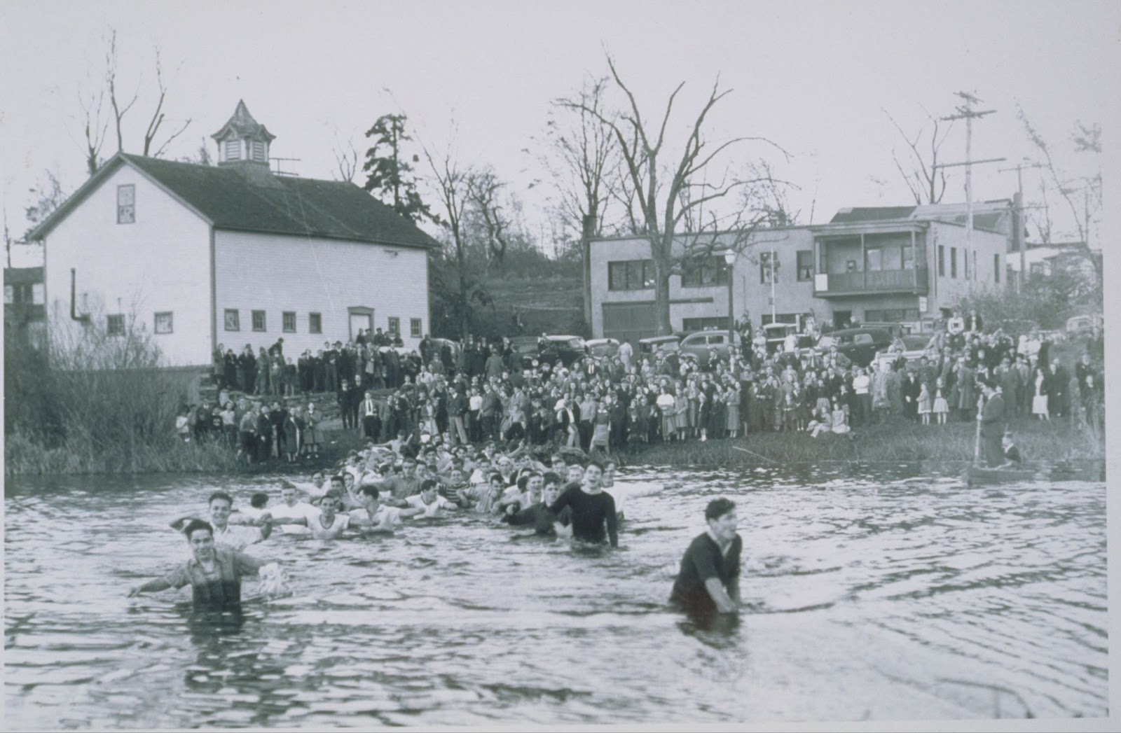 Many men wading fully-clothed and waist-deep in a body of water, with a crowd looking on