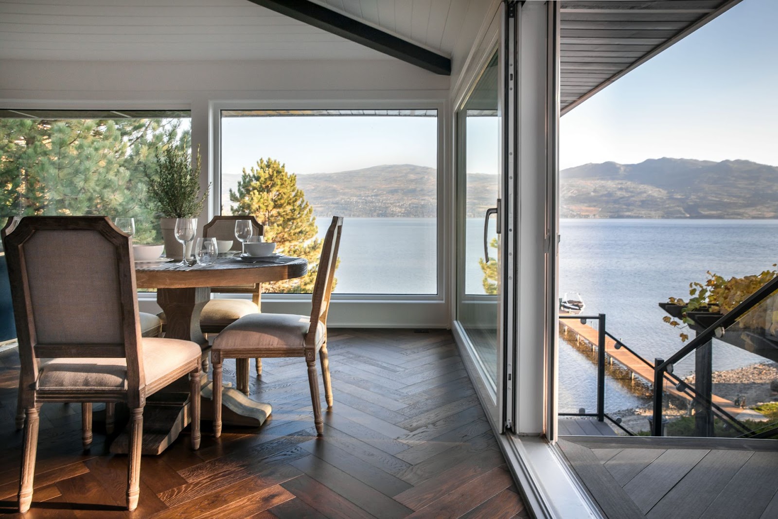 Image of a house looking out at Okanagan Lake with steps going down to the dock on the right side. A table and chairs are set up on the left with large picture windows. 