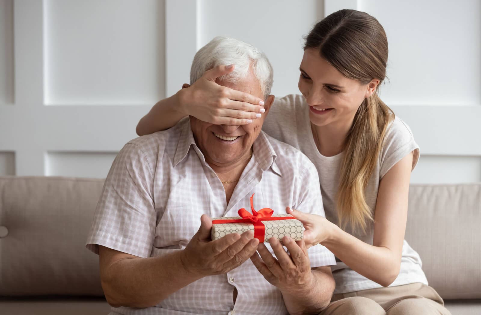 An adult child covering their senior parent's eyes while smiling and preparing to open a small gift.