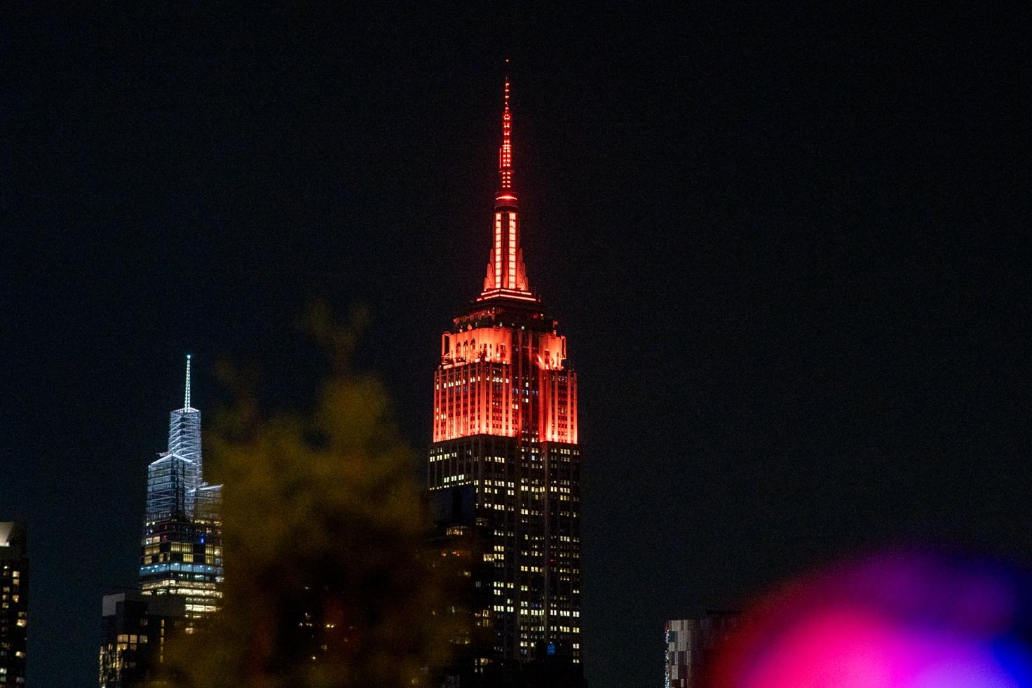 A tall building with red lights with Empire State Building in the background Description automatically generated