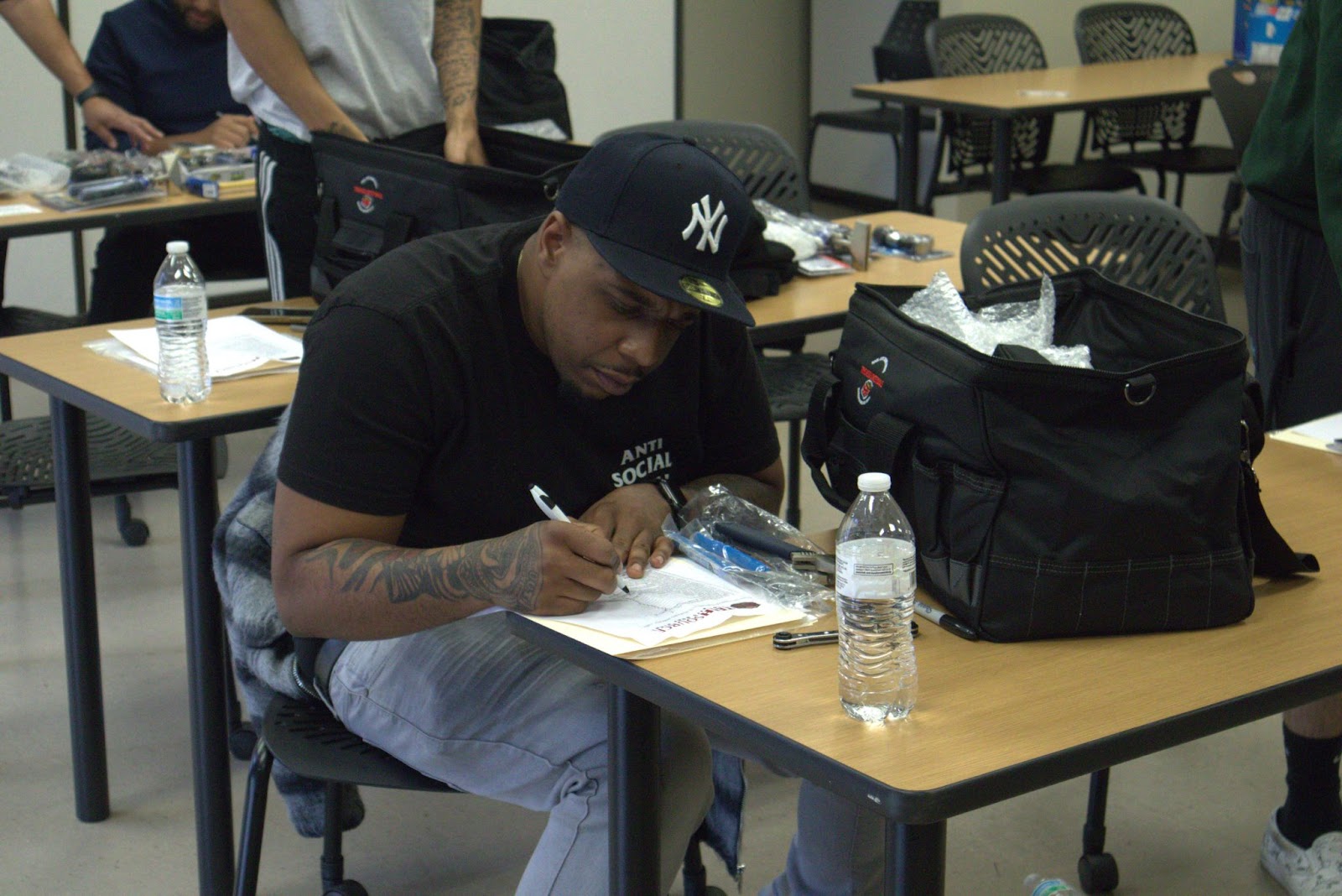 A student seated at a desk is focused on completing paperwork in a classroom setting. The desk holds tools, a water bottle, and a black tool bag, emphasizing a hands-on training environment. Other students are visible in the background, engaging in similar tasks, highlighting a collaborative and productive learning atmosphere.