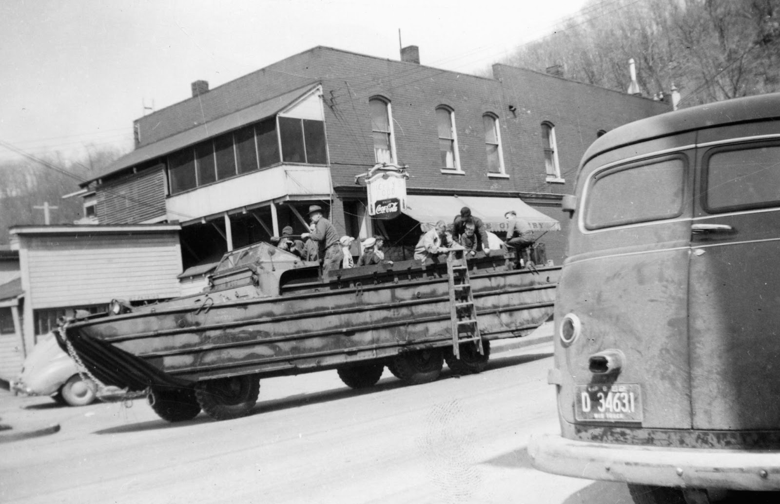 A group of men stand on a boat on wheels in the middle of a street. Cars are parked along both sides of the street with buildings in the background. 