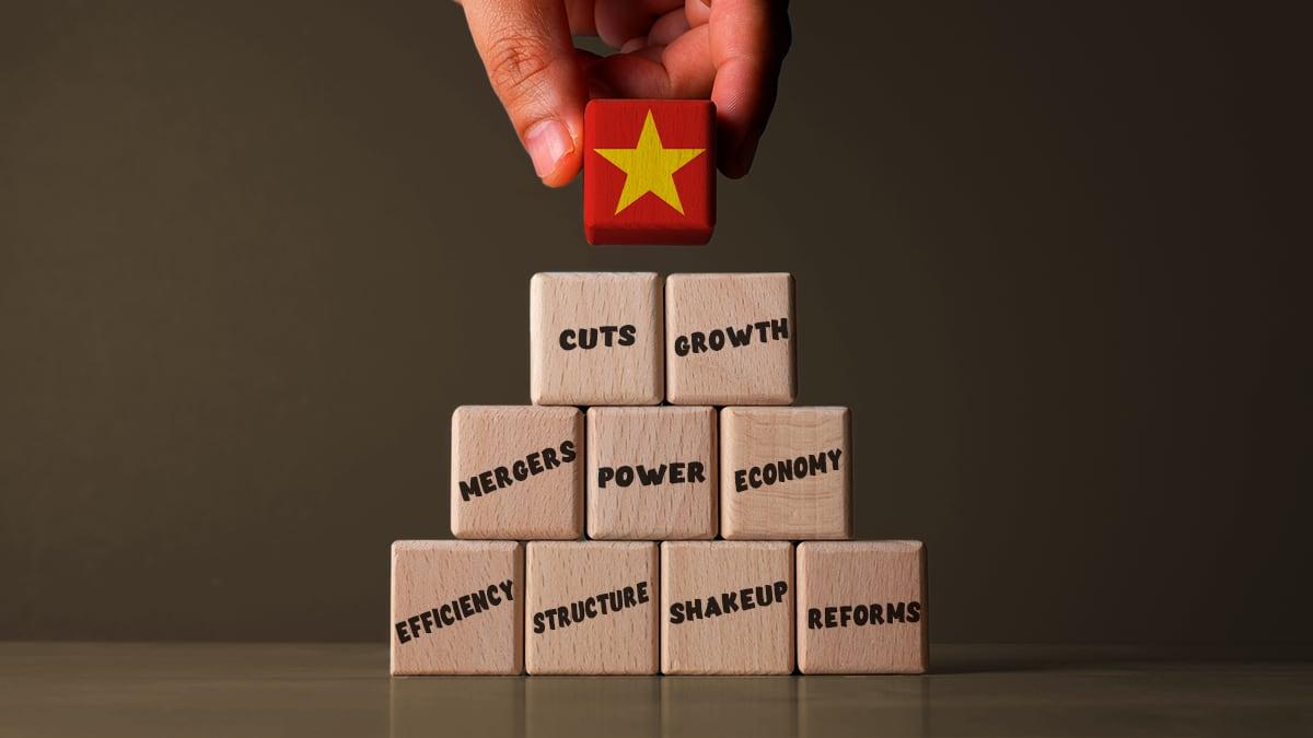 A hand places a block with the Vietnamese star on top of a stack of other blocks with the words cuts, growth, mergers, power, economy, efficiency, structure, shakeup and reforms written on them.