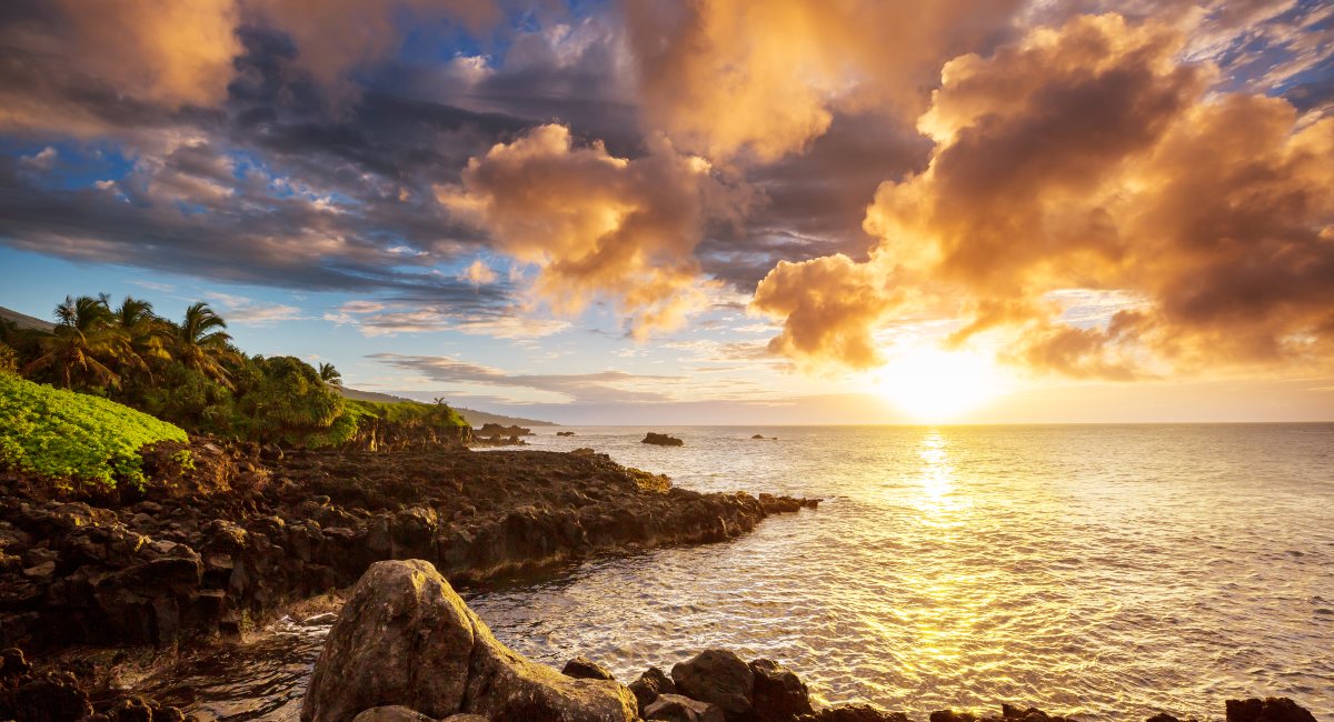 Breathtaking sunset over Maui's rocky coastline with golden clouds and sparkling ocean waters, highlighting lush tropical vegetation and dramatic cloud formations.