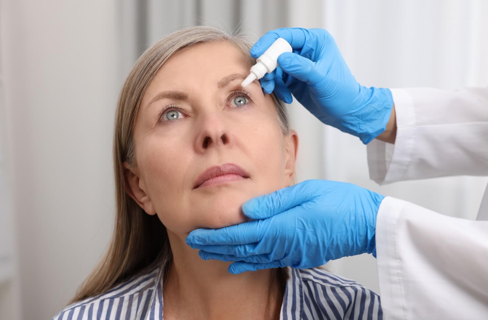 A person looks upward as a healthcare professional wearing blue gloves carefully administers eye drops.