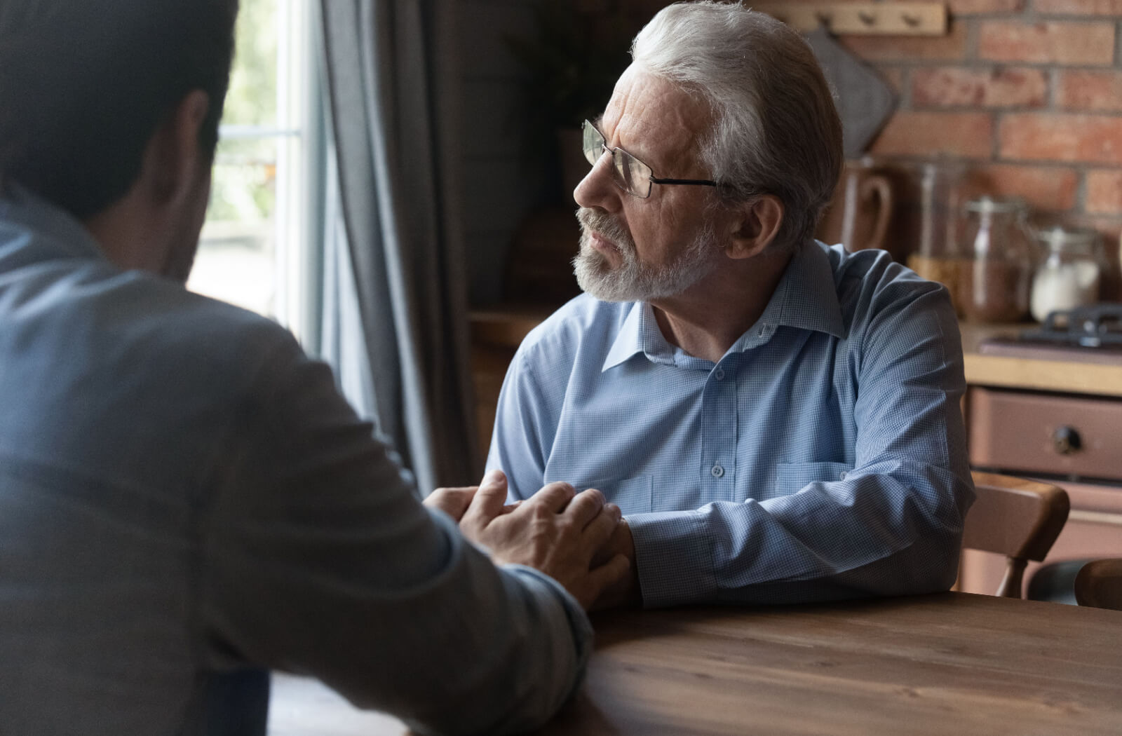 An older adult with memory loss looking out the window of their kitchen while their adult child holds their hand and reassures them.