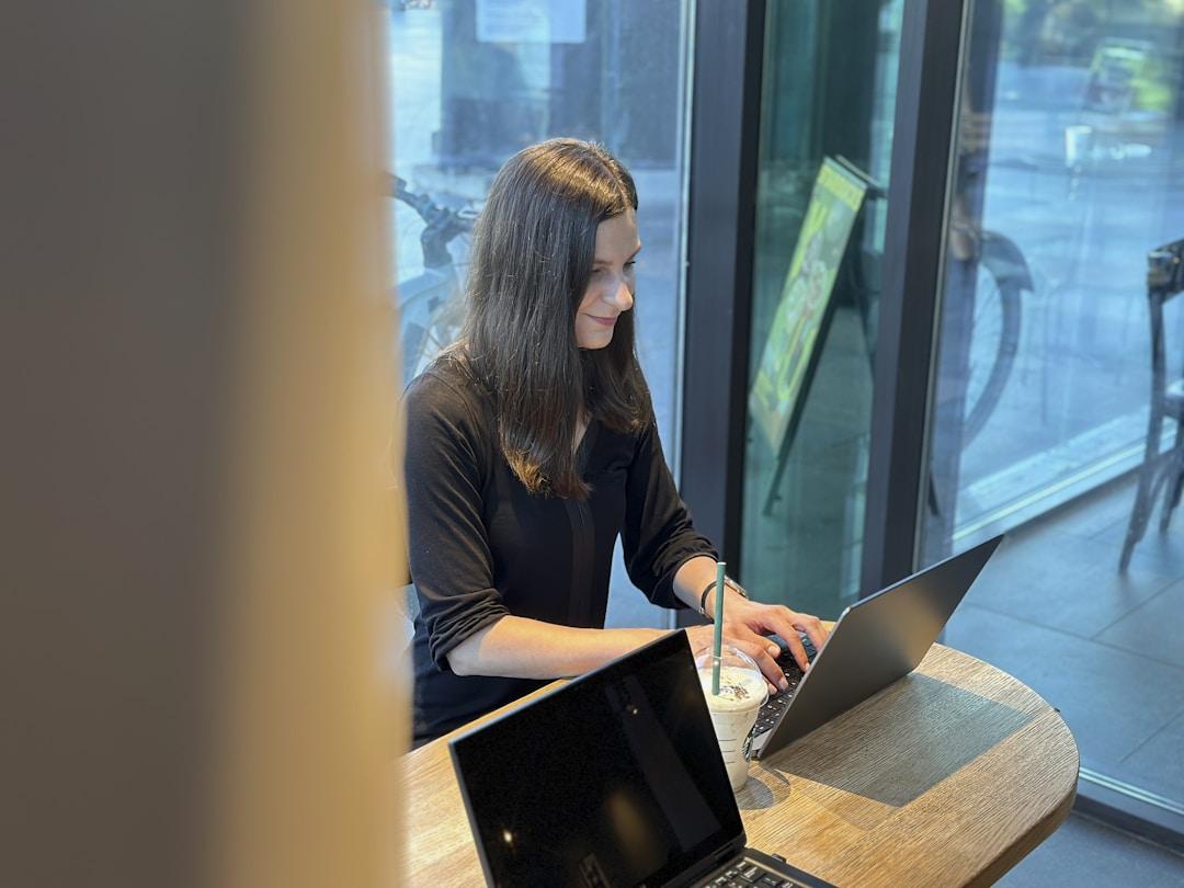 Individual working on a laptop at a wooden table in a well-lit indoor space, with a beverage cup and a second electronic device beside the laptop.