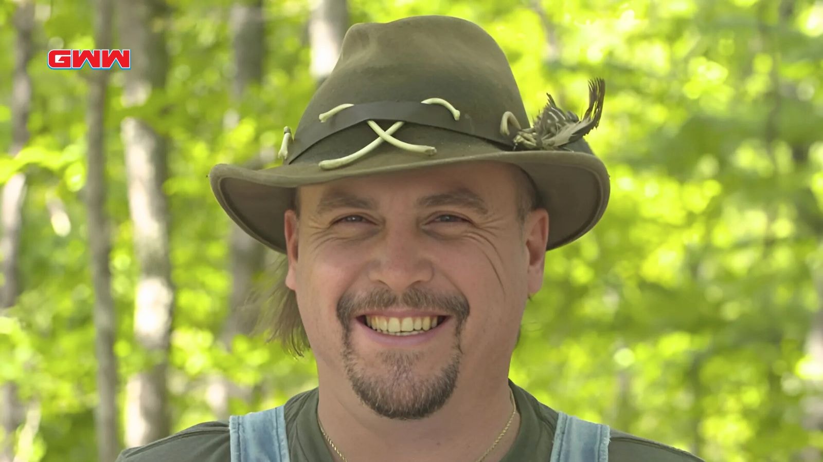 Mike with a feathered hat smiling outdoors in a wooded area.