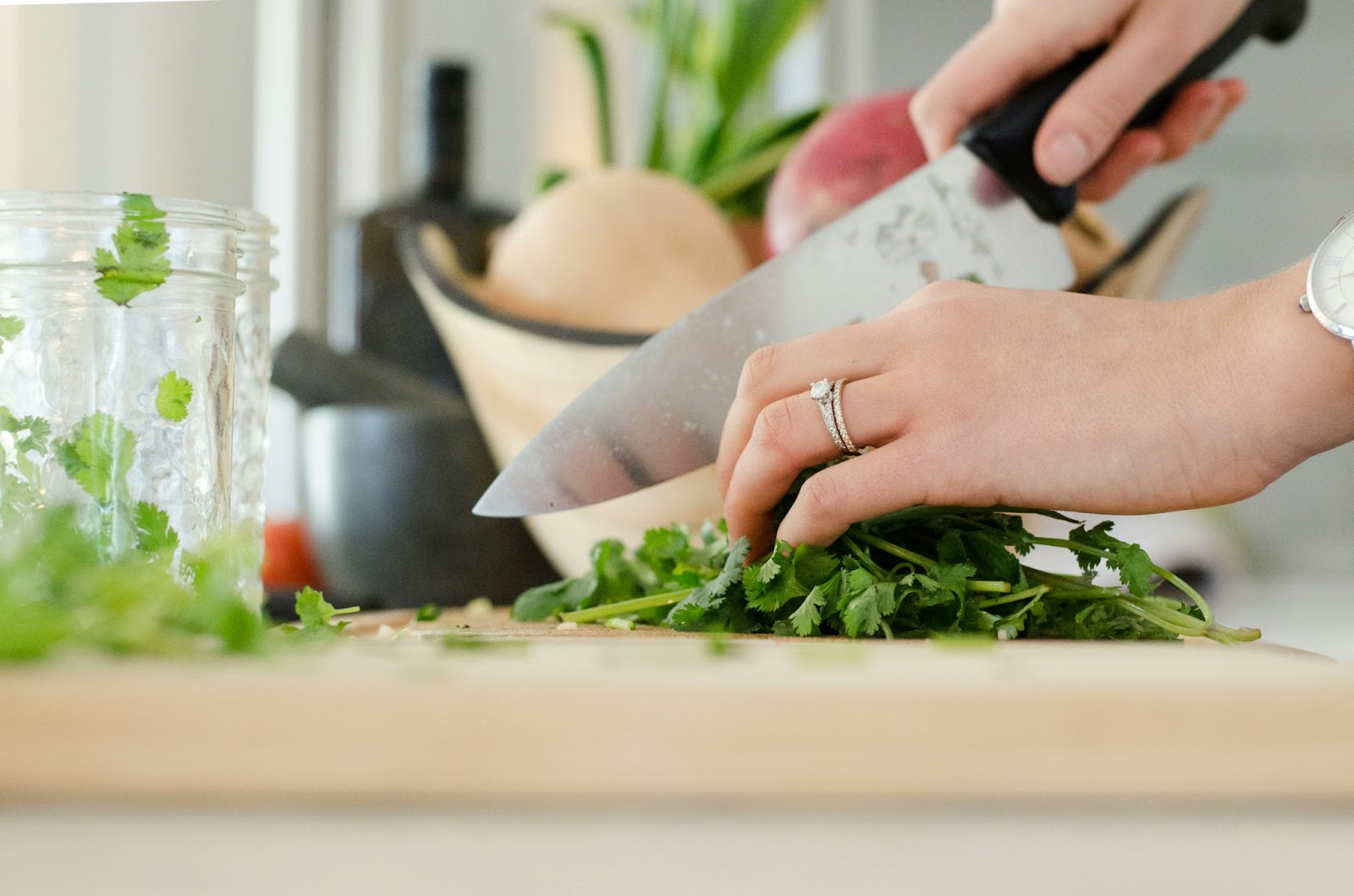 Close-up of a woman with a wedding ring chopping cilantro with a large silver knife and placing it into a glass jar.