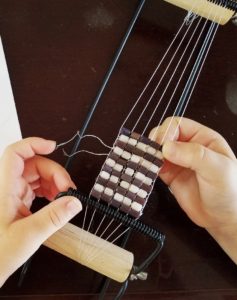 A child's hands weave a thread through purple and white beads on a bead loom