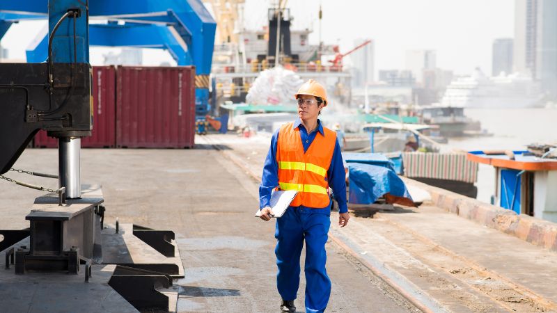 Un ouvrier portant un casque et un gilet de sécurité marche sur un quai très fréquenté avec des documents ; des conteneurs d'expédition, des grues et des bâtiments de la ville sont visibles.