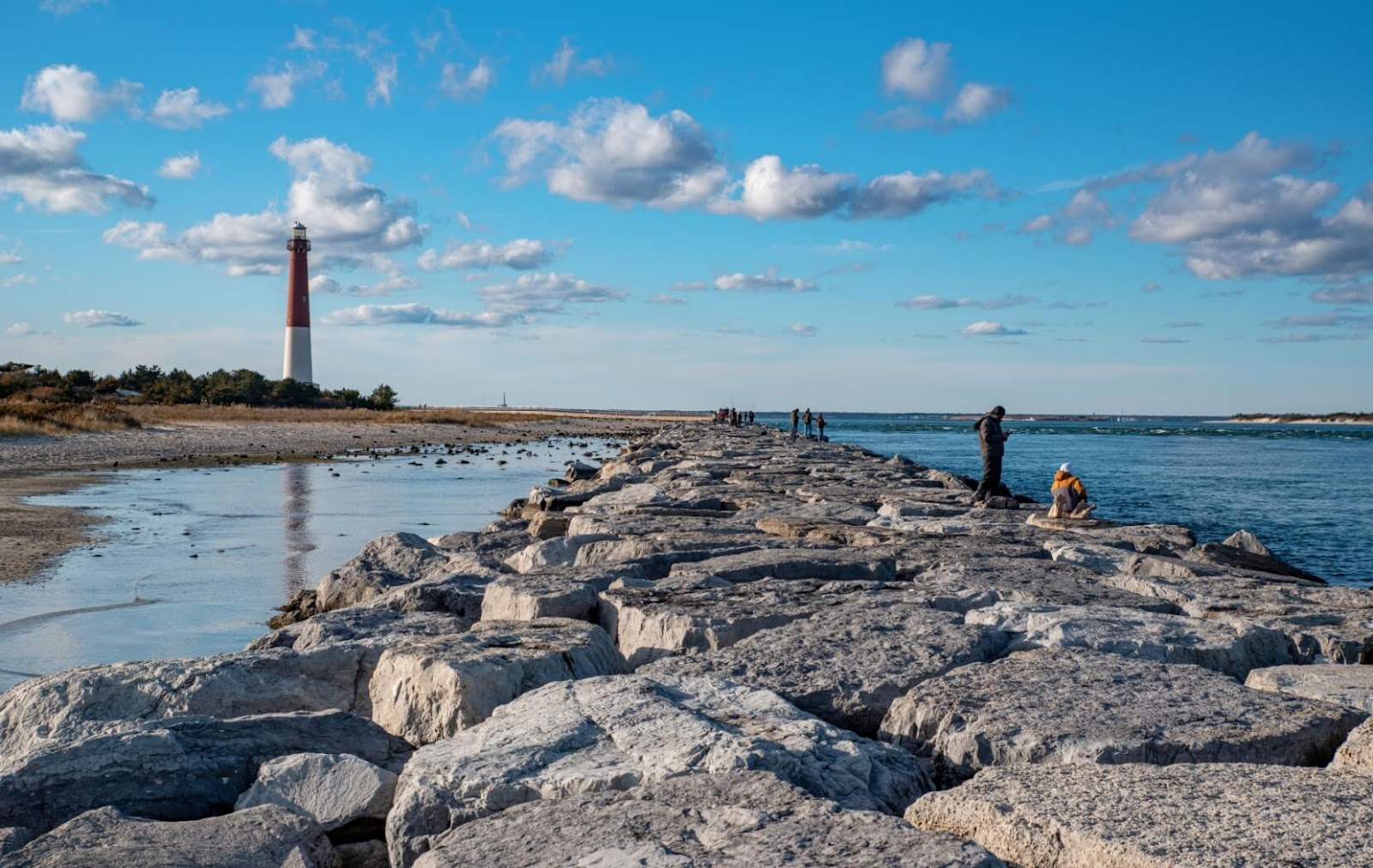 Visitors walk across the rocks of Barnegat Lighthouse State Park while the Barnegat Lighthouse stands in the distance.