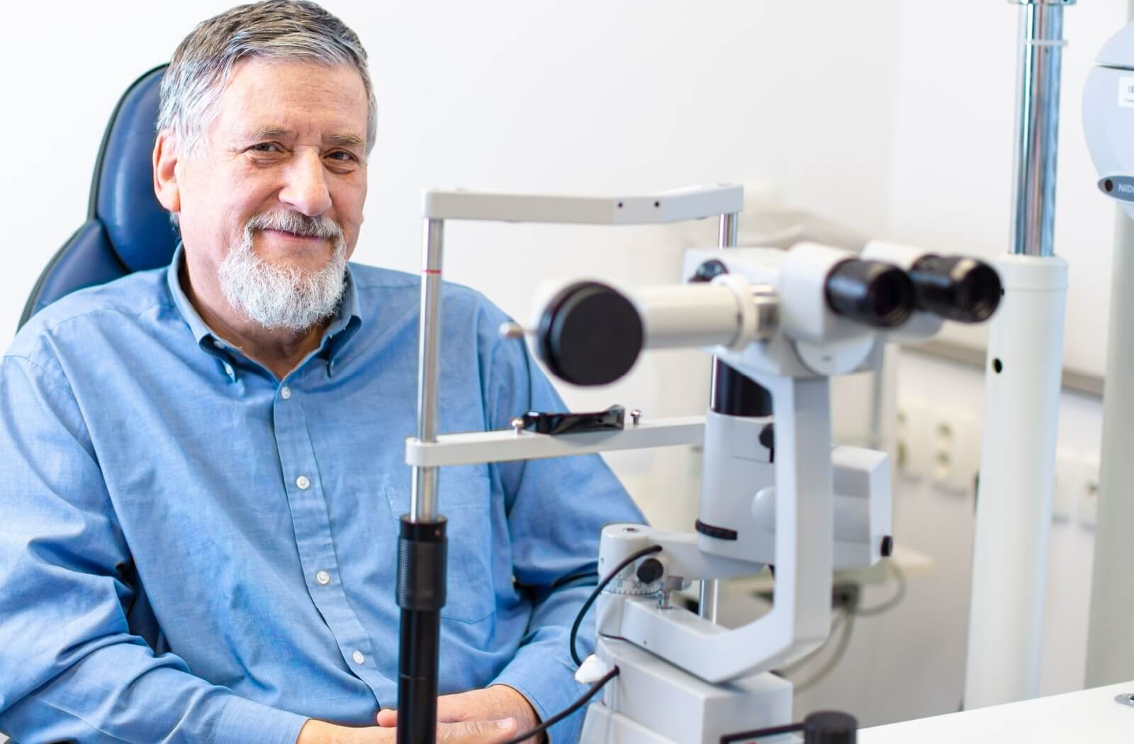 A smiling older man undergoing an eye exam, seated behind some diagnostic equipment.