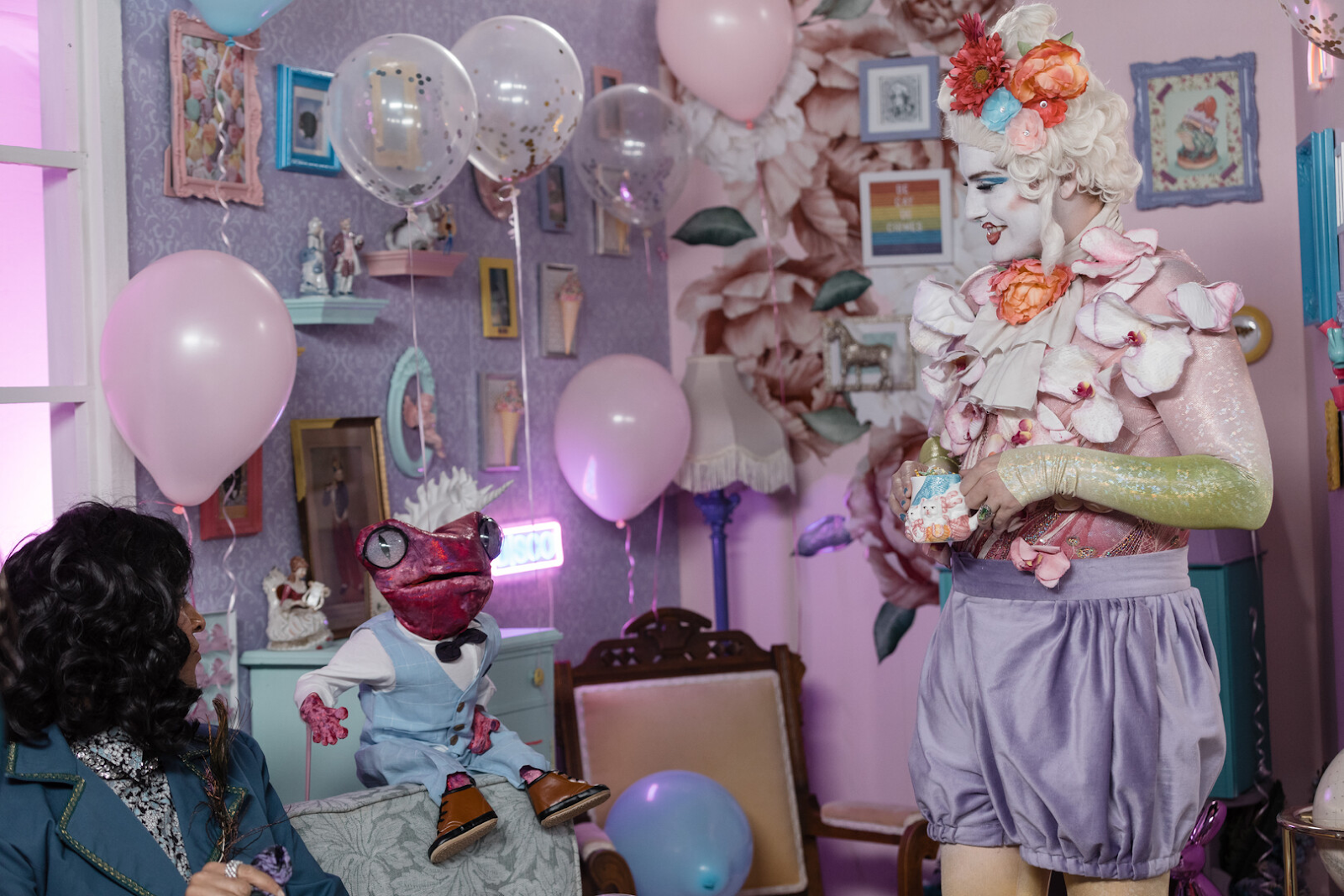 A pastel playhouse filled with knick-knacks, balloons, a drag character dressed like an 18th century nobleman, a guest, and a weird little puppet