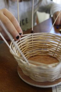 A child's hands are threading beads onto the upright frame of a basket in the process of being woven