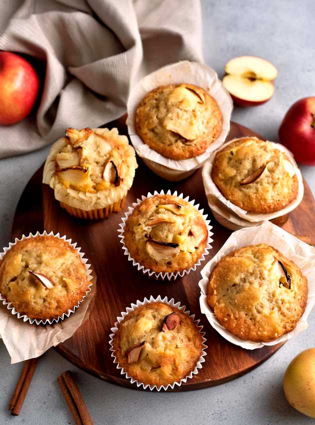 Apple muffins with cinnamon on a wooden serving board, surrounded by fresh apples.