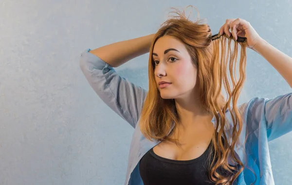 A woman with light brown hair attaching a clip-in hair extension, standing against a light blue wall, wearing a black tank top and an open shirt.