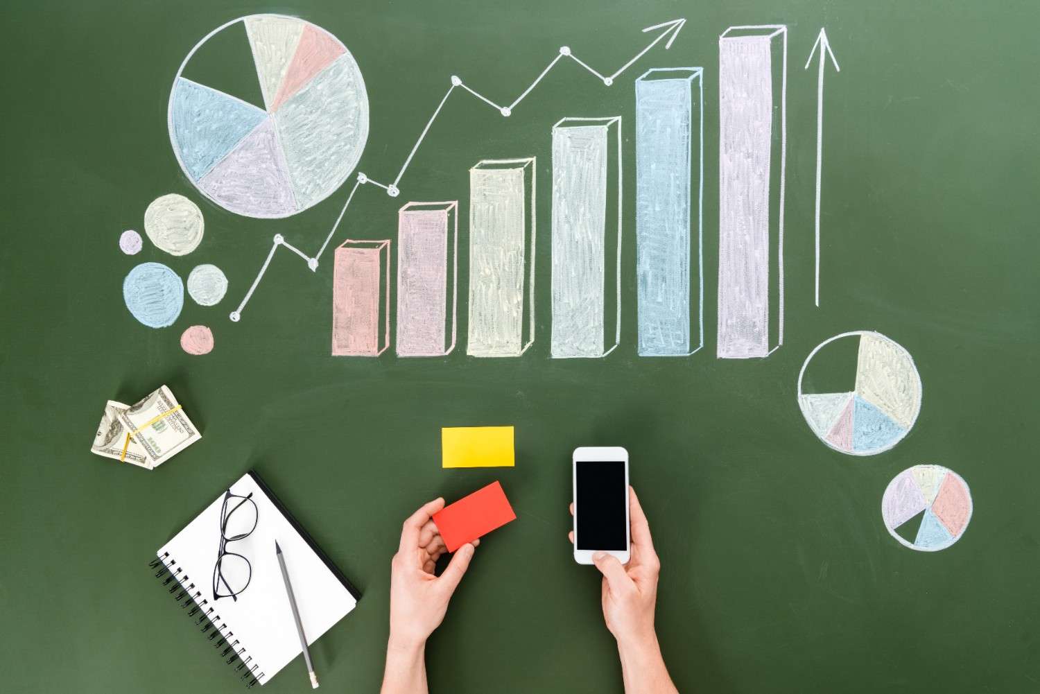 A top view of a workspace with hands holding a smartphone and a red card on a green chalkboard background. The chalkboard features colorful graphs, pie charts, and a line chart representing financial or business data. A notebook, glasses, a pen, and cash are placed nearby