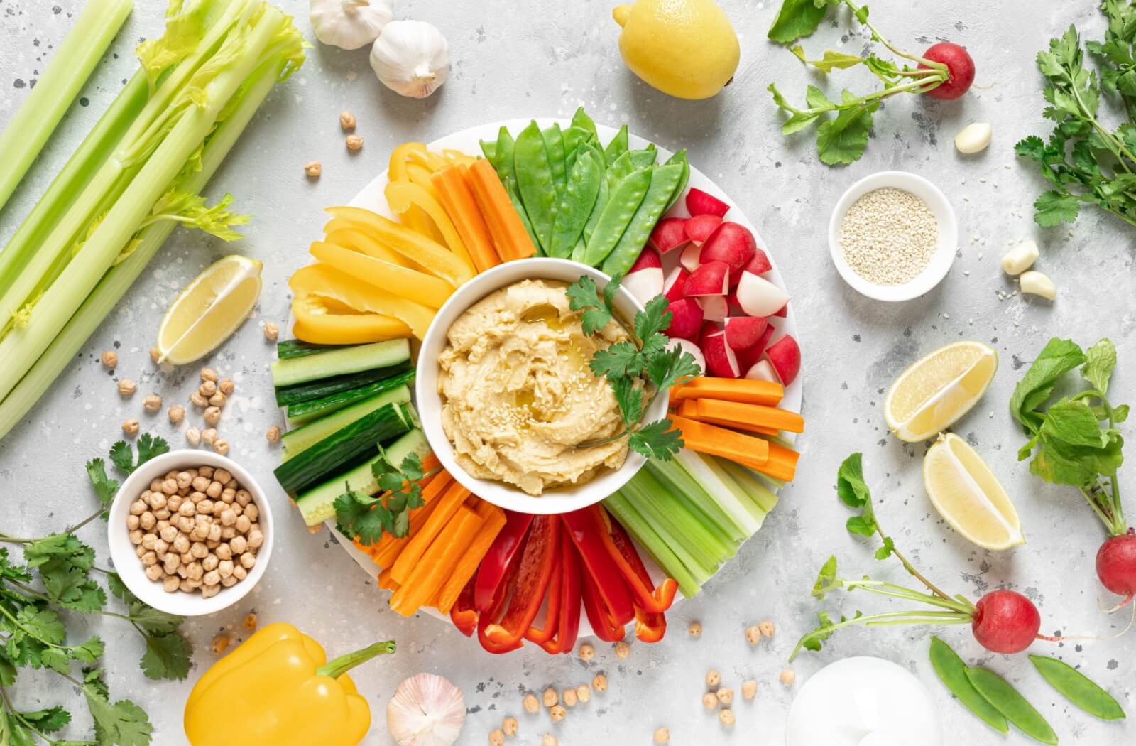  A plate of protein-rich hummus with assorted veggies surrounded by a display of celery, lemons, and other ingredients.