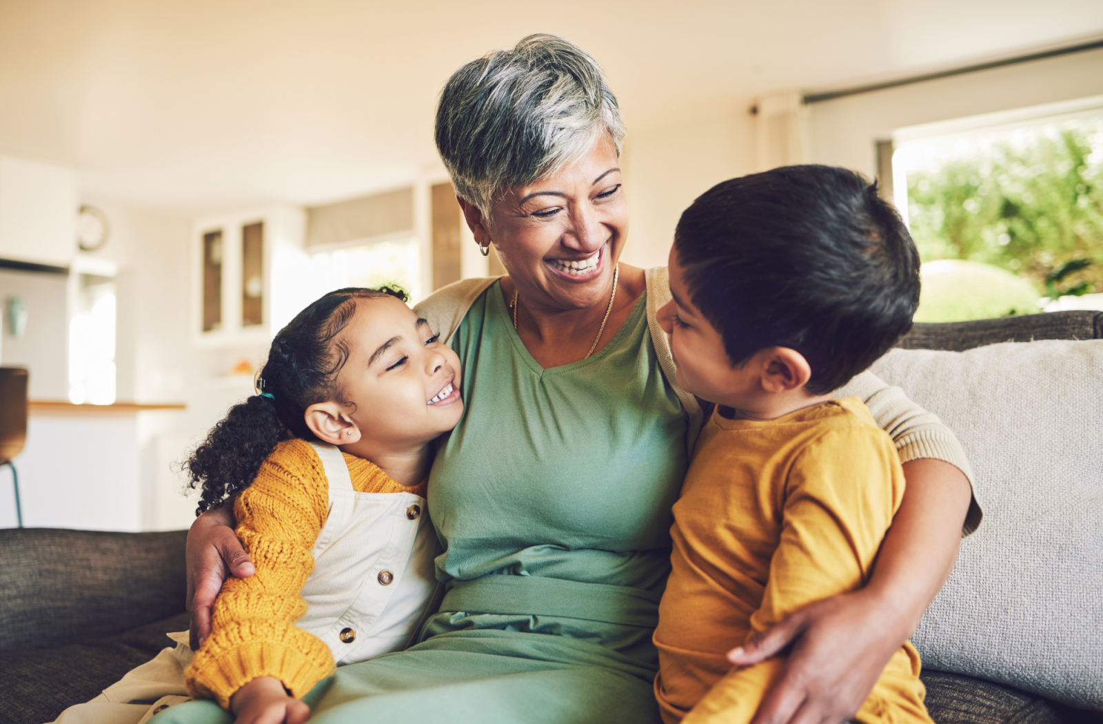 A grandparent holds 2 young grandchildren close on the couch, laughing together.
