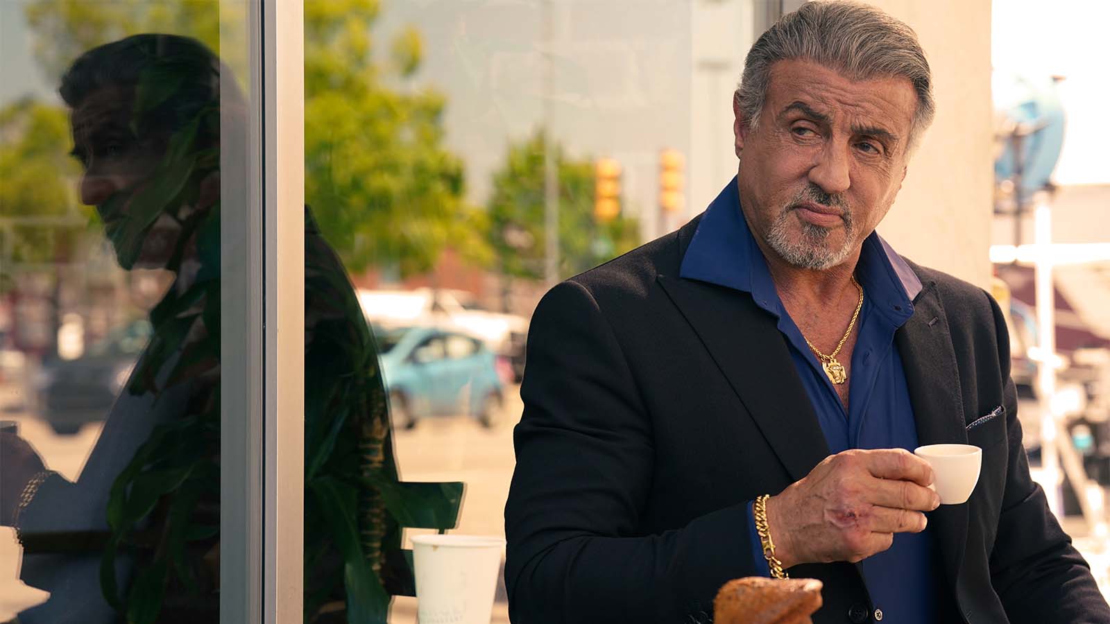 Sylvester Stallone en un traje oscuro con una camisa azul y accesorios dorados, sosteniendo una taza de café mientras mira hacia un lado. Reflejo en una ventana cercana, con un entorno urbano de fondo. Expresión seria y actitud relajada del actor.