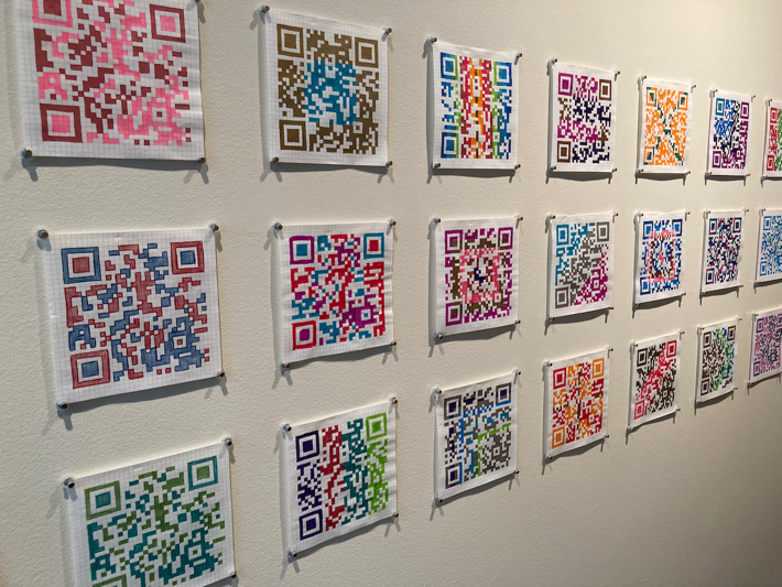 QR codes made by hand hang on a wall. Each is a different set of two, three, or more colors.