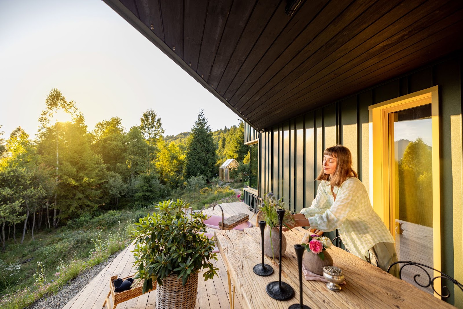  A woman arranges a plant in a vase on a wooden table on a deck overlooking a lush garden, enjoying the tranquil morning.