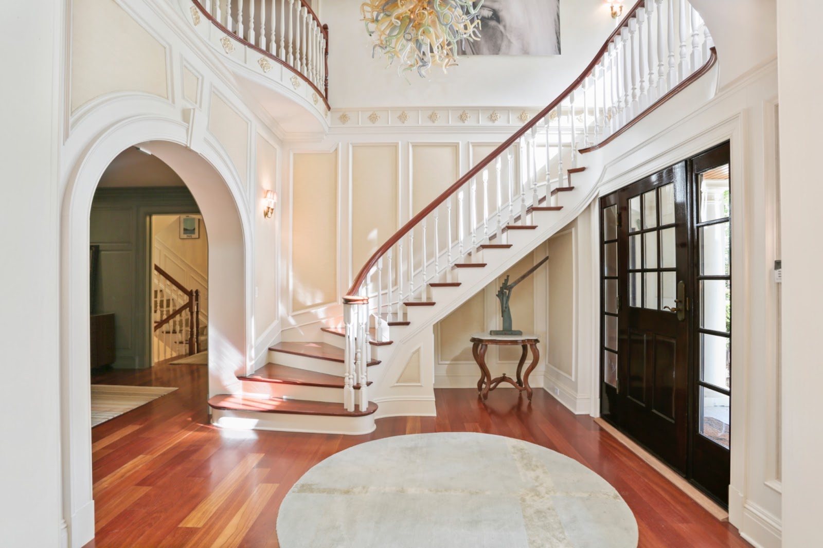 A grand two-story foyer entryway features a sweeping staircase with table and sculpture in alcove beneath the stairs, Murano-style glass sculpture chandelier and large mural art above the stairs.