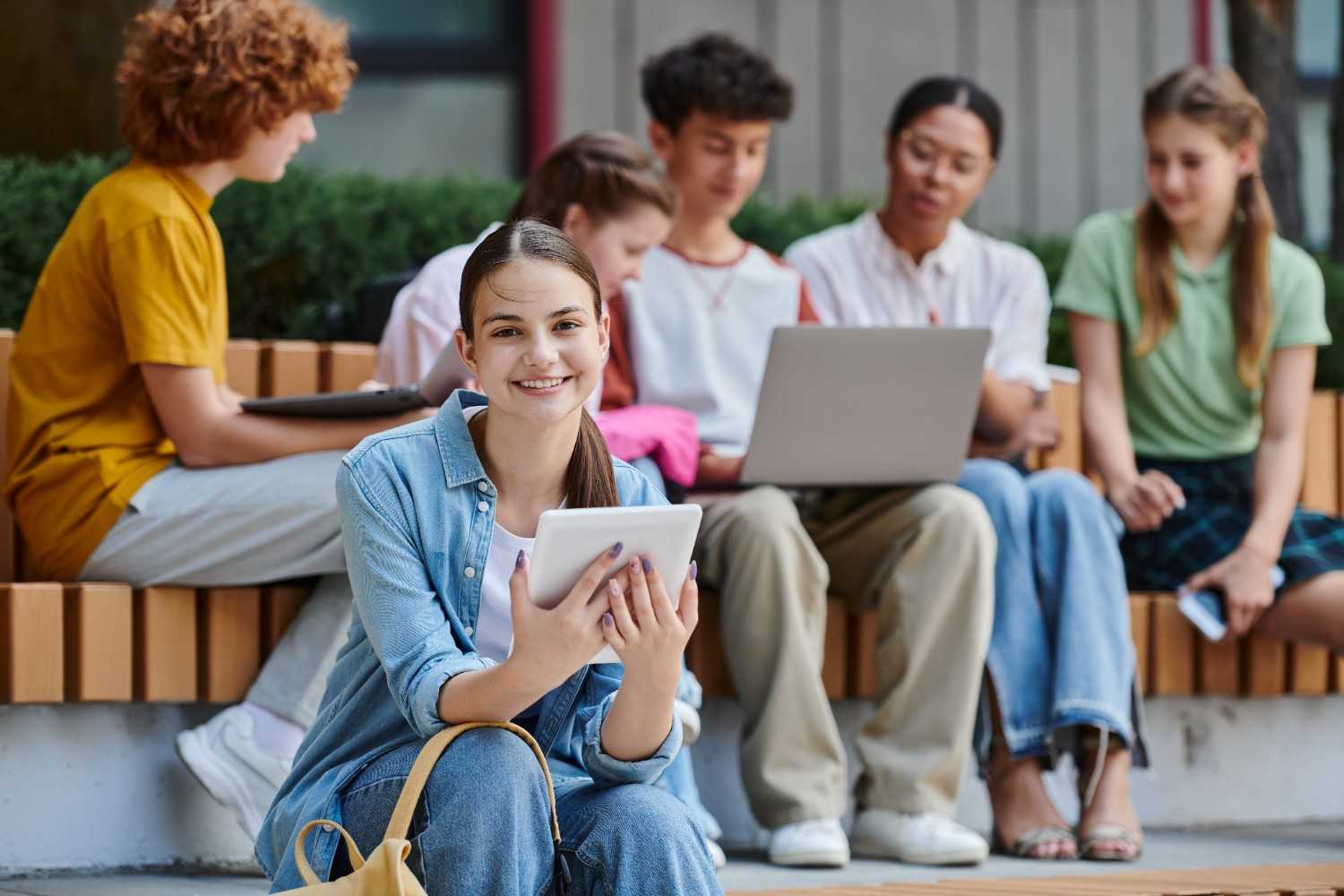 A group of diverse students sitting outdoors on a bench, engaging in collaborative work with laptops and tablets. A smiling girl in the foreground holds a digital tablet, wearing a denim shirt and looking directly at the camera