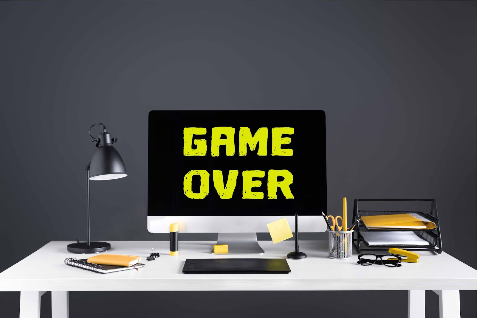 (A computer on a desk displays the text "GAME OVER" in bold yellow letters, hinting at entertaining virtual icebreaker games. The desk also features a lamp, stationery, and stacked documents.)