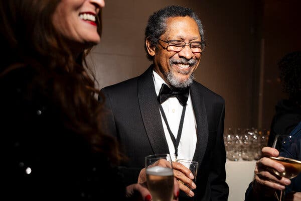 This is a photo of the author Percival Everett. He is wearing a tux with a white shirt and bow tie, and there is a medal around his neck. He is holding a glass in one hand and smiling broadly.