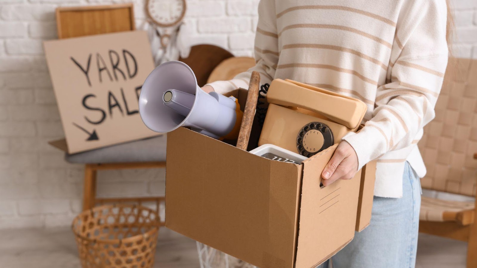 A person preparing for a yard sale, holding a cardboard box filled with assorted items like a telephone, a megaphone, and various household goods, with a "Yard Sale" sign in the background