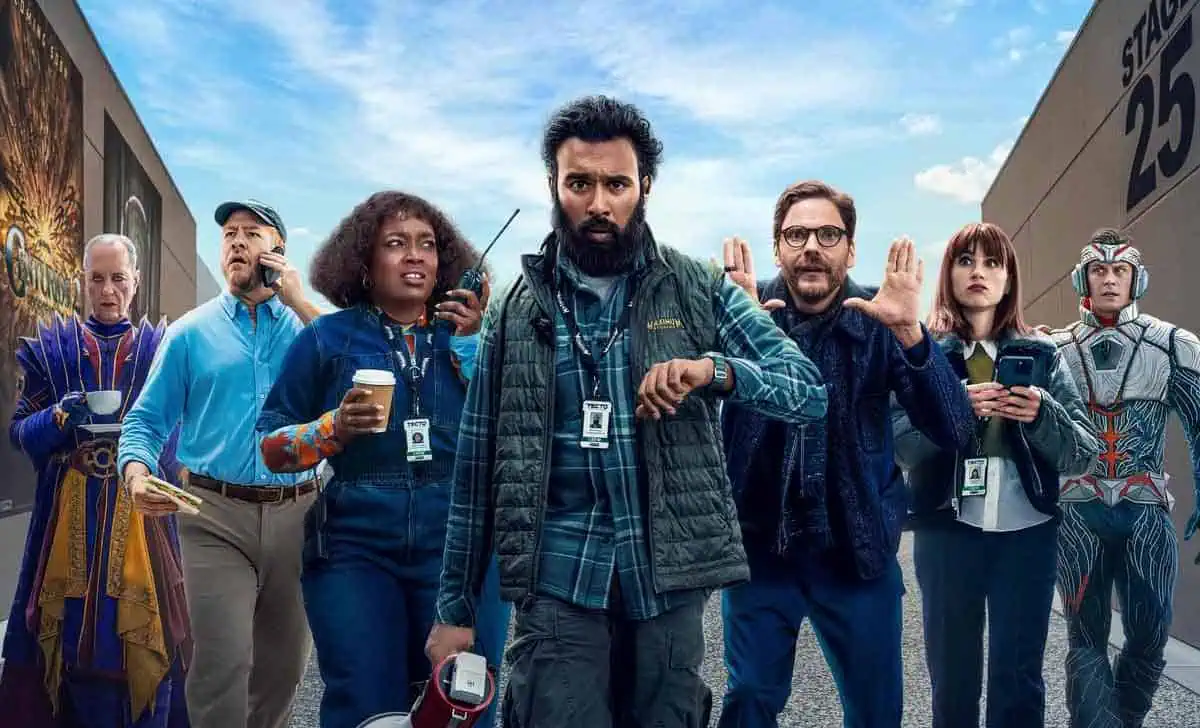 Grupo del equipo de producción de La Franquicia caminando por un estudio al aire libre. Daniel Brühl y Himesh Patel lideran al equipo, con ambos haciendo gestos de acción. A su alrededor, el elenco está vestido con atuendos casuales, y se destacan los fondos que recrean un set cinematográfico.