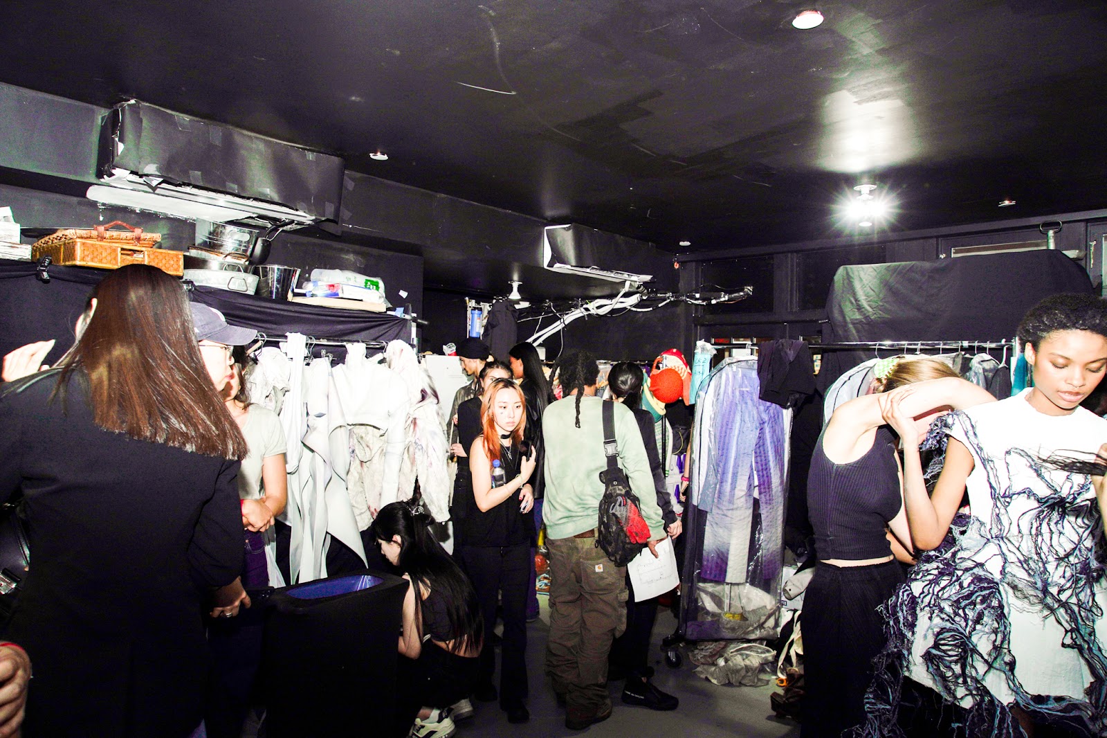 A chaotic backstage environment shows designers, models, and crew preparing for a fashion show, with racks of clothing and various garments strewn around the room.