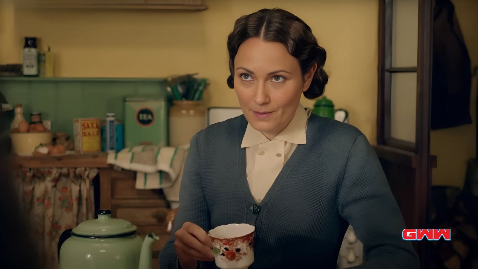 Anna Madeley as Mrs. Audrey Hall holding a tea cup in the kitchen.