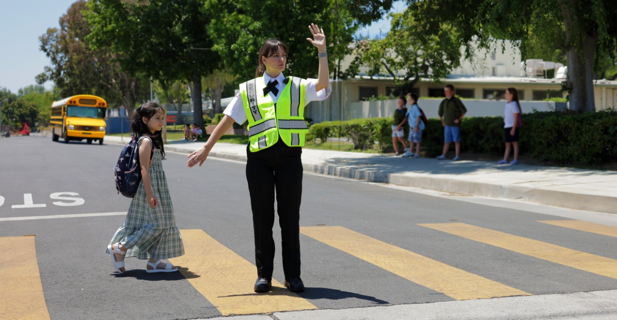 (A Citiguard security officer helps a young student safely cross the street near a school zone, with other children waiting on the sidewalk.)