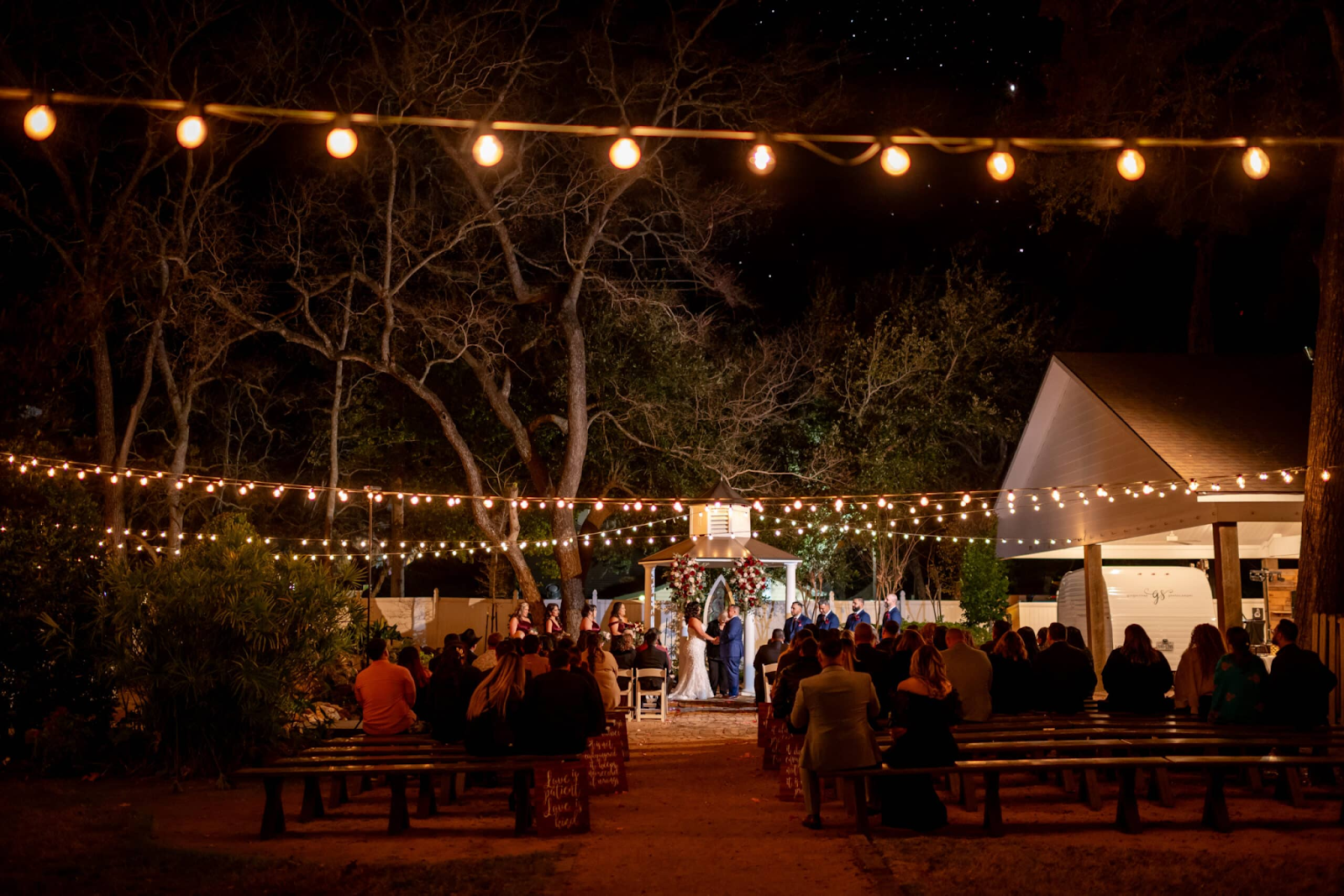 Under the magical glow of twinkling string lights, a night wedding ceremony unfolds at the gazebo, one of the many perfect wedding ceremony locations at Butler's Courtyard in League City, Texas. Guests are comfortably seated on custom wedding benches that perfectly complement the venue's style, adding to the enchanting and elegant ambiance of the event.