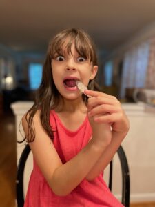 A Caucasian girl with brown hair and wide eyes takes a bite out of a piece of white rock candy. 