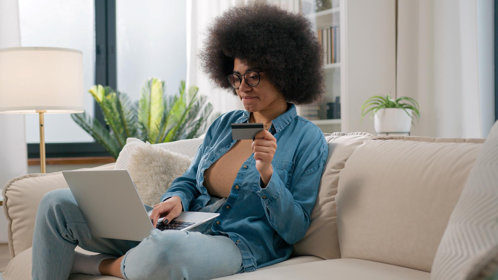 A lady with a full afro hair sitting on a couch with a credit card on one hand and a laptop on her lap wondering why thd/cbna on her credit report