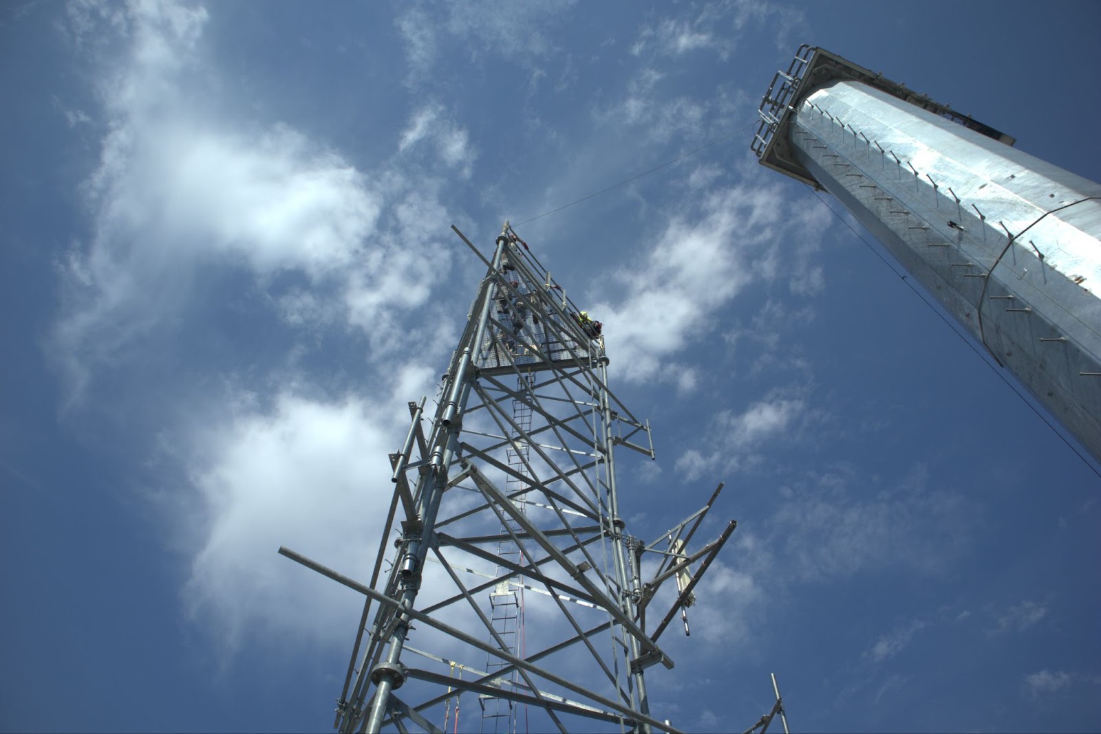 A telecommunications tower technician wearing a helmet, harness, and safety gear is working on a metal tower structure. The technician is carefully secured to the tower, focusing on a task while positioned high above the ground. The bright sky serves as a backdrop, highlighting the technician’s elevated work environment and emphasizing the importance of safety measures in this field.