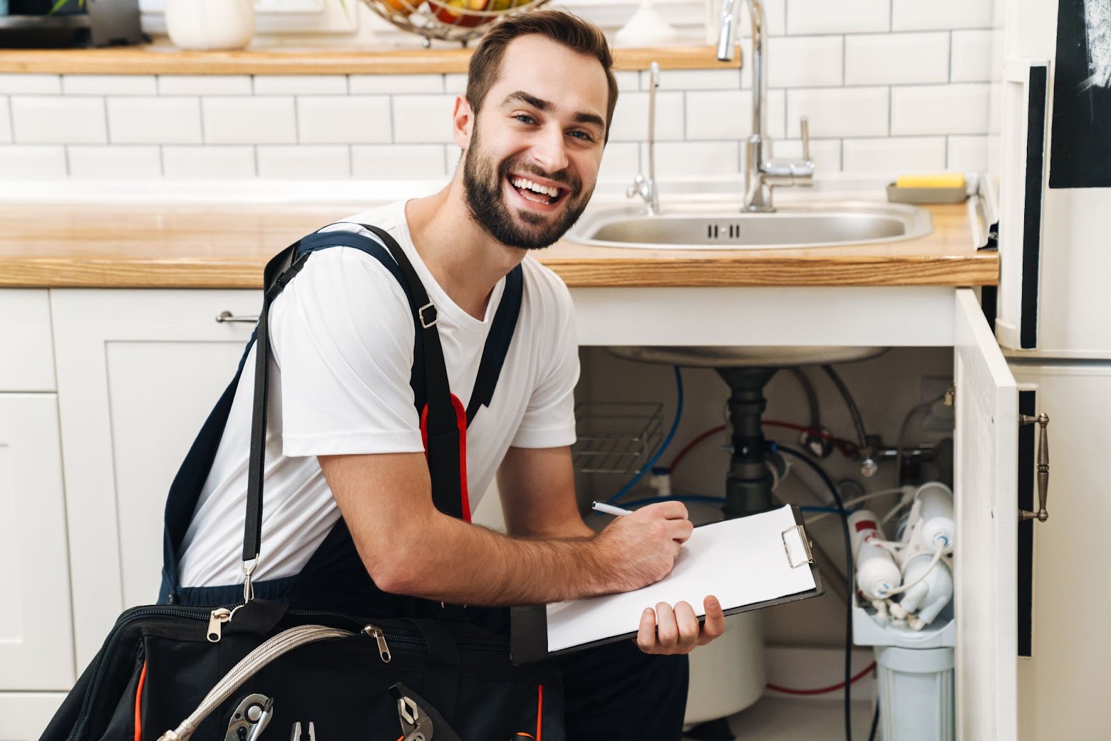 Smiling plumber with tools next to a sink writing on a clipboard. 