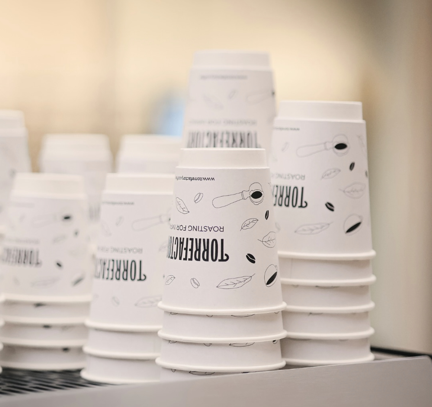 Stack of branded white paper cups with a simple black coffee bean and leaf design, neatly arranged on a counter in a coffee shop.