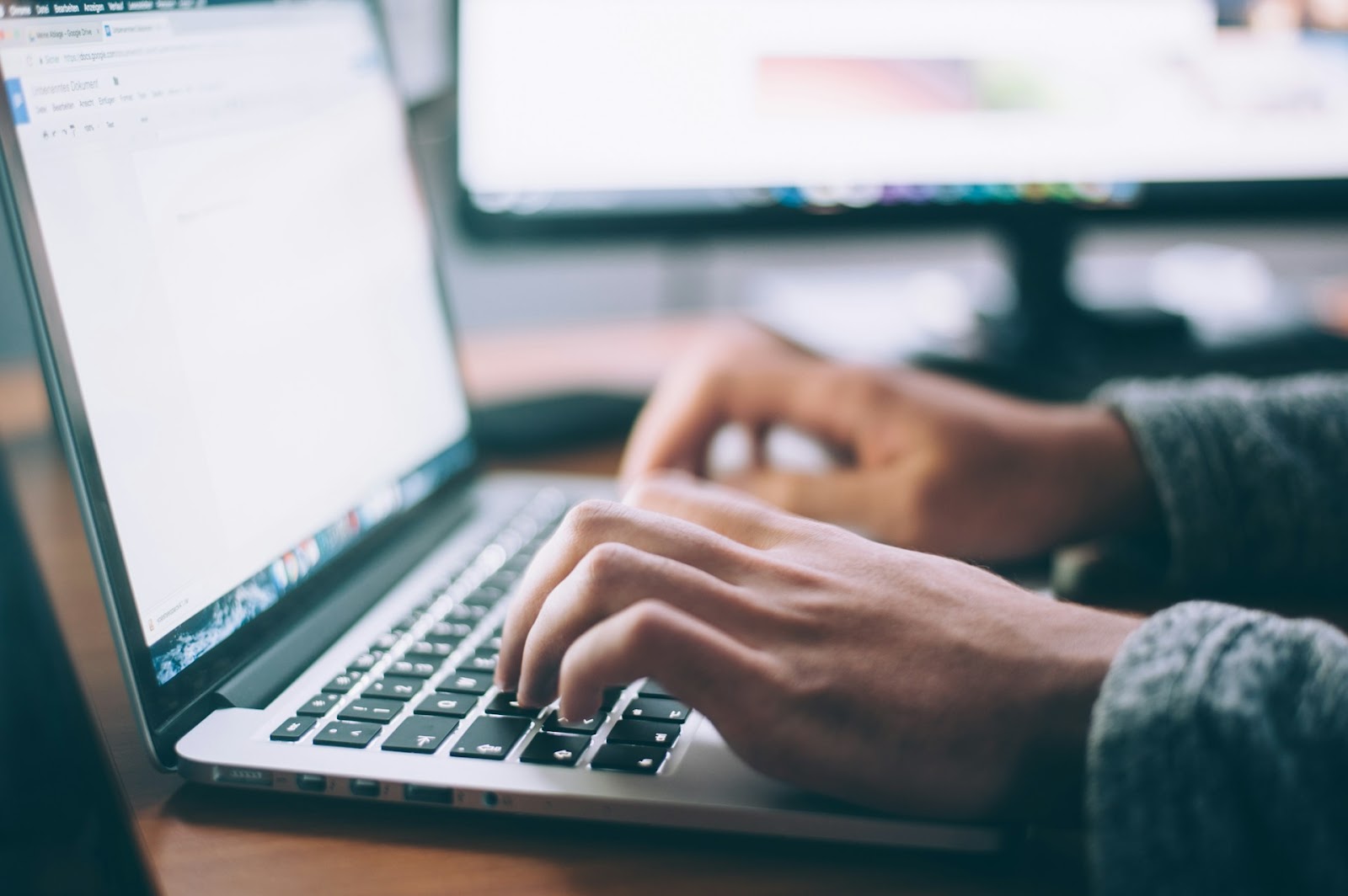 Close-up view of a person's hands typing on a laptop, focusing on how to create online courses, with a blurred background featuring another computer screen.